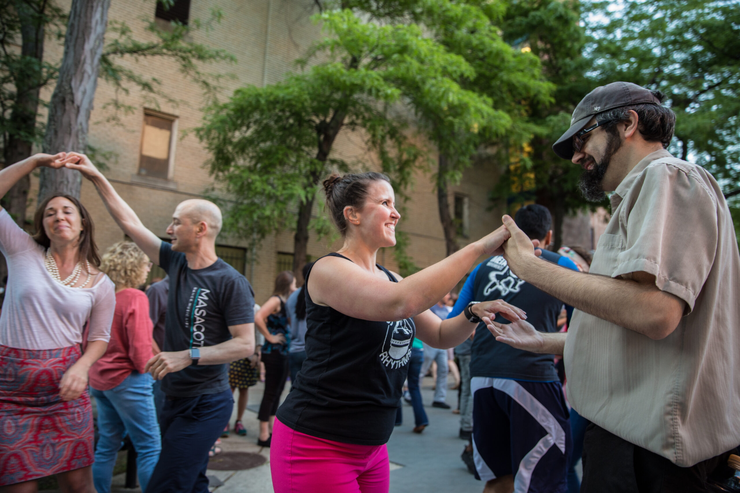A group of people dancing at Salsa in the Park. Dancers are partnered, with one pair joining their hands and smiling while another spins nearby.