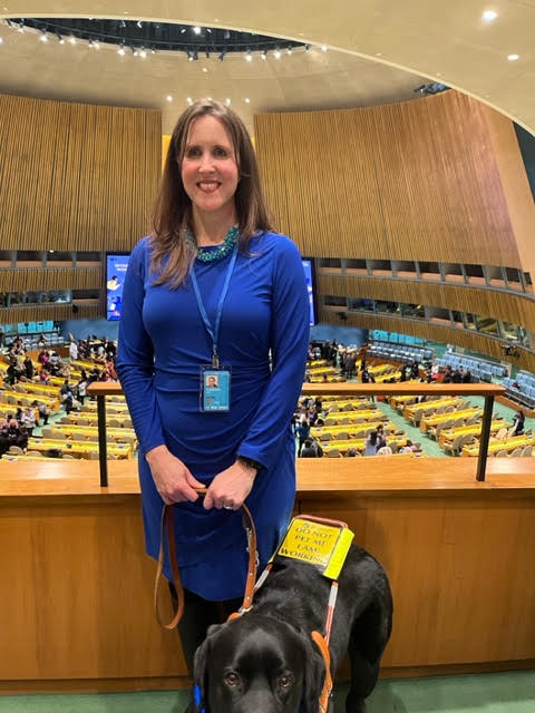 Kerry and her guide dog Miles, a black lab, smile at the camera. They are standing in the upper area of an auditorium at the United Nations General Assembly in New York City.