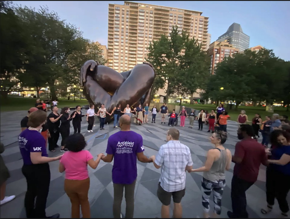 Kerry’s salsa class stands in a circle holding hands in front of the “Embrace” statue at The Boston Commons. She is signing to the group wearing a clear mask with an interpreter doing ProTactile on her back.