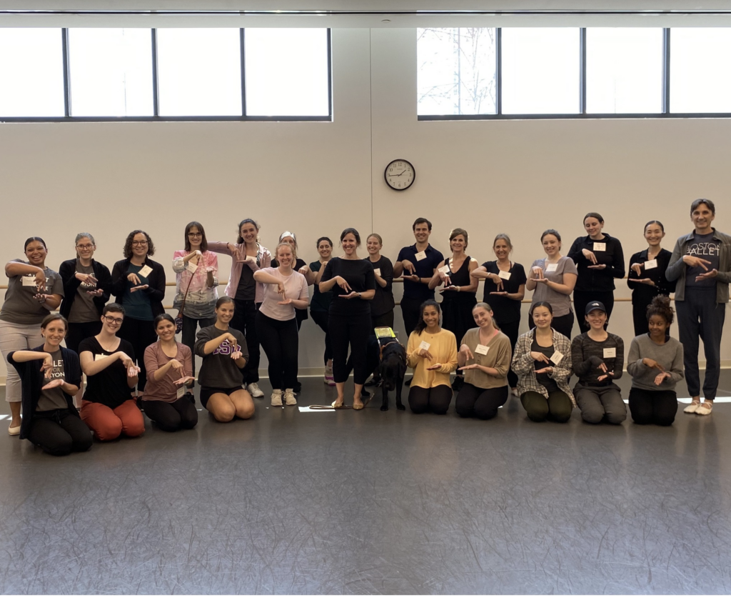 Kerry, Executive Director of Silent Rhythms, is surrounded by a group of Boston Ballet students in two rows. Miles, Kerry’s black lab guide dog, is standing to her right wearing his harness. Kerry and all the students are signing DANCE and smiling at the camera.