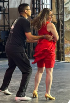Kerry in a red dress and gold shoes dancing with a partner in black shirt, black pants.